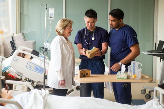 Health students in scrubs look at a clipboard with a professor in a lab coat