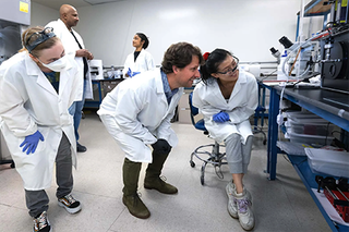 Professor surrounded by students crouches and looks at machinery