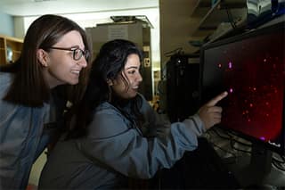 Two people in lab gear point at a red screen